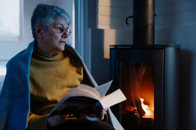 una anciana se sienta cómodamente cubierta con una bufanda cerca de una chimenea caliente y lee su libro favorito