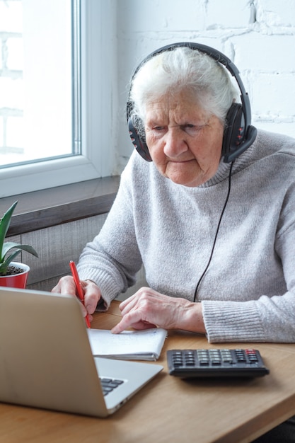 Una anciana se sienta en una mesa frente a una computadora portátil con auriculares y escribe en una libreta.