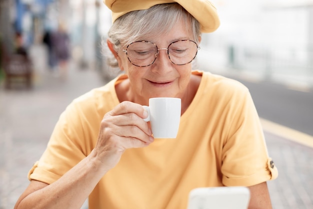 Una anciana sonriente y atractiva vestida de amarillo disfrutando de un mensaje de lectura de una taza de café exprés en el teléfono
