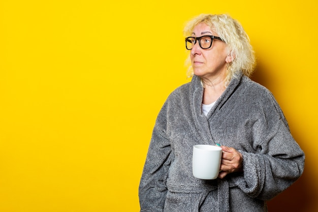 Anciana sonriente en bata de baño gris sosteniendo una taza con café mirando hacia el lado