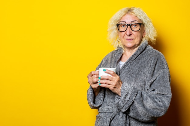 Anciana sonriente en bata de baño gris sosteniendo una taza con café