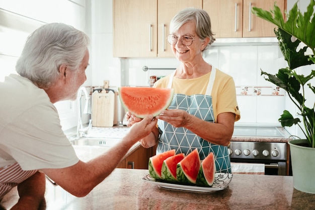 Una anciana sonriente en la cocina de su casa ofreciendo a su esposo una rebanada de sandía fresca de temporada jubilados y un concepto de estilo de vida saludable