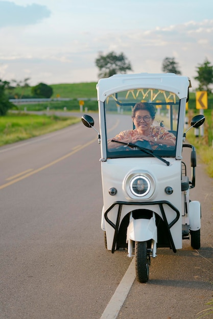 Una anciana sonriente conduciendo un triciclo eléctrico en la carretera con paisajes de arrozales