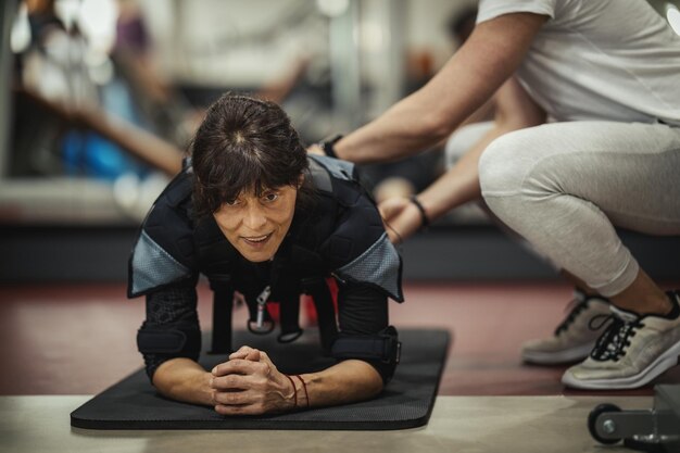Foto una anciana sonriente está haciendo ejercicios de tabla con un entrenador personal en el gimnasio, vestida con un traje negro con un simulador electrónico ems para estimular sus músculos.