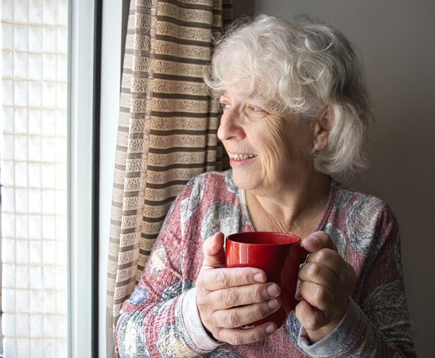 Una anciana sonriente de pie con una taza de café en las manos y mirando  por la ventana | Foto Premium