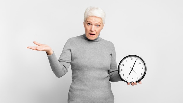 Una anciana sorprendida sosteniendo un reloj sobre un fondo gris