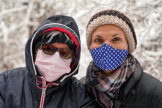 Anciana y su hija menor vestidas de invierno, ambas con mascarilla de virus, fondo borroso de árboles cubiertos de nieve