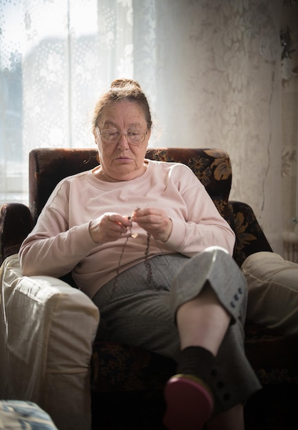 Una anciana con suéter beige sentada en el sillón y tejiendo