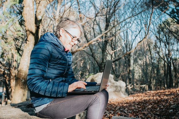 Una anciana súper feliz y sonriente usando su laptop para bloguear en línea durante una caminata, aprendiendo a usar tecnología y actividades durante la tercera edad. Cuidado de la salud para personas mayores y naturaleza deporte abuela