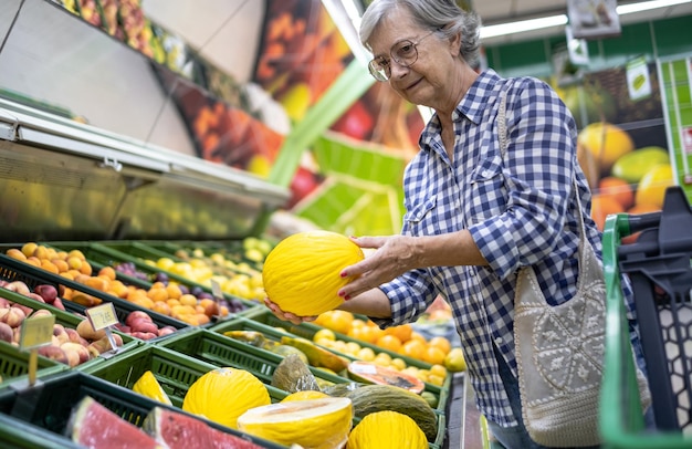 Anciana en el supermercado eligiendo fruta fresca sosteniendo un melón amarillo en su mano Cestas de frutas de temporada en el fondo