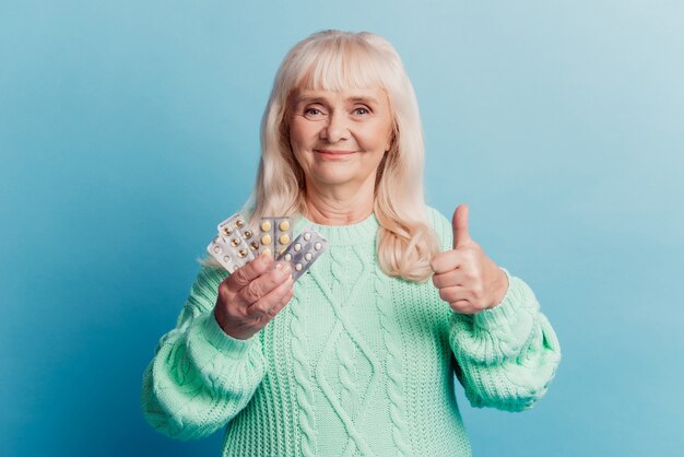 Anciana con tabletas de retención de productos médicos muestran el pulgar hacia arriba sobre fondo azul.