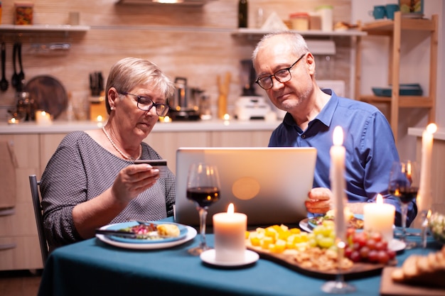 Anciana con tarjeta de crédito mientras usa la computadora portátil con su esposo para ir de compras. Ancianos sentados a la mesa, navegando, usando la tecnología, internet, celebrando su aniversario.