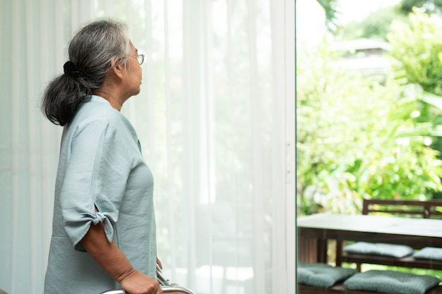 Una anciana triste usa andador para pararse frente a las ventanas y mirar afuera y sentirse sola