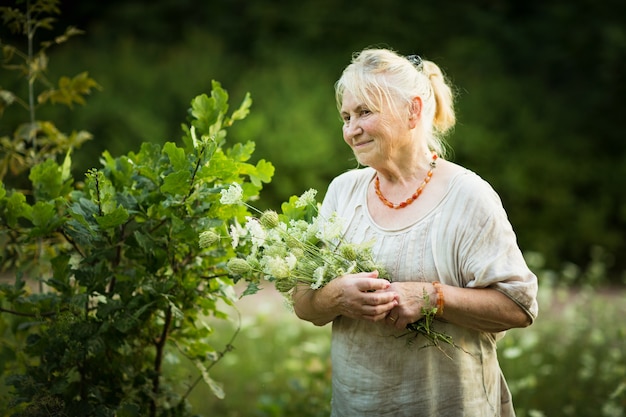 Anciana en vestido blanco vintage camina por el jardín con un ramo de flores de campo y sonrisas