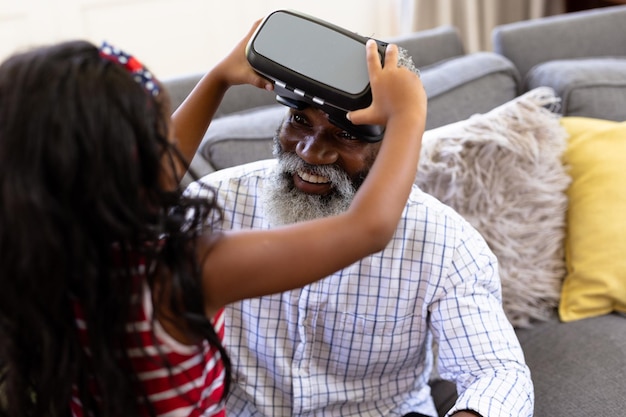 Un anciano afroamericano y su nieta disfrutan juntos de su tiempo en casa, sentados en un sofá, usando un auricular VR