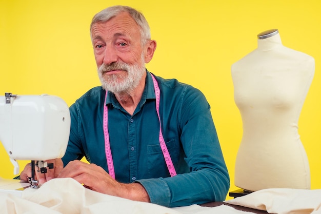 Un anciano agradable trabajando en un estudio de sastrería haciendo ropa en una máquina de coser, fondo amarillo.