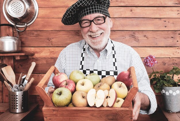 Un anciano alegre y atractivo sonriente sosteniendo una cesta llena de manzanas de varias calidades y colores