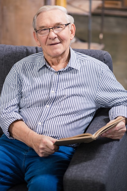 Anciano alegre sonriendo mientras lee un libro en casa