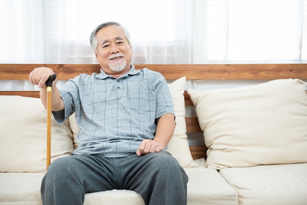 Anciano anciano senior sentado, descansando sus manos sobre un bastón de madera sentado en el sofá en la sala de estar en casa después de la jubilación.