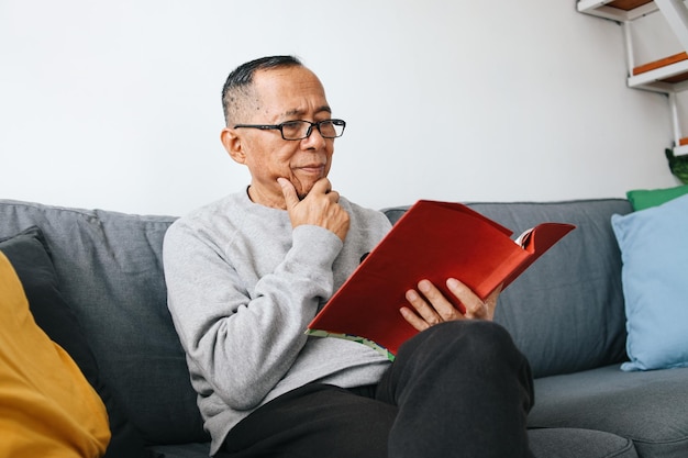 Un anciano asiático con gafas leyendo un libro en el sofá.