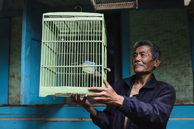 Anciano asiático sosteniendo una jaula de pájaro verde fuera de una casa de madera tradicional