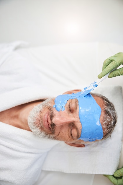 Anciano de aspecto relajado tiene el rostro cubierto con una espuma azul en un salón de spa