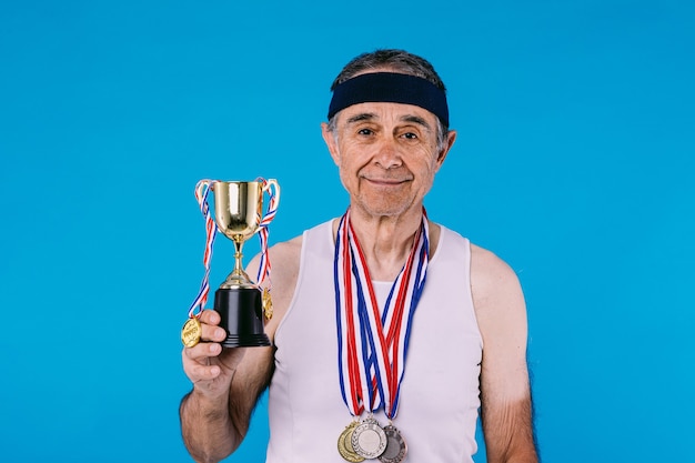 Anciano atleta con marcas de sol en los brazos, con tres medallas en el cuello sosteniendo un trofeo con cintas colgando, sobre fondo azul.