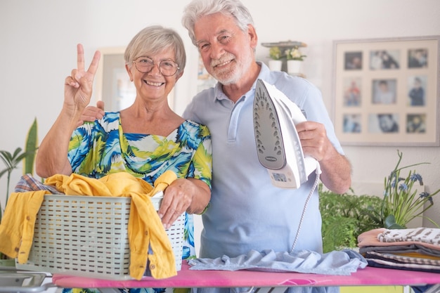 El anciano ayuda con las tareas domésticas planchando la ropa en la tabla de planchar mientras su feliz esposa lleva una canasta con otra ropa