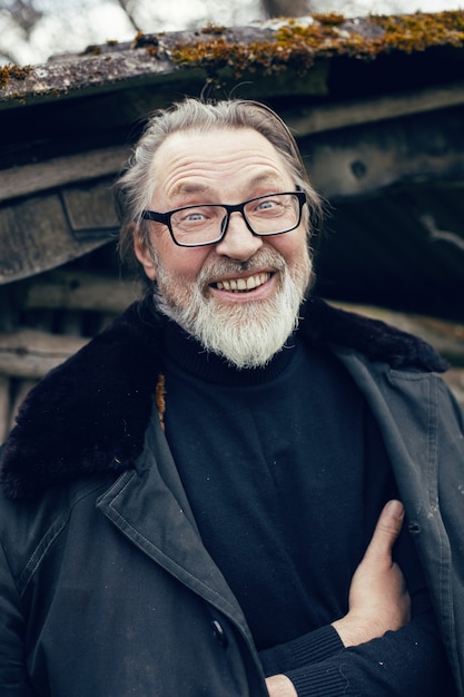 Anciano con barba se encuentra en la aldea cerca de un cobertizo de madera con un abrigo de piel de oveja y gafas