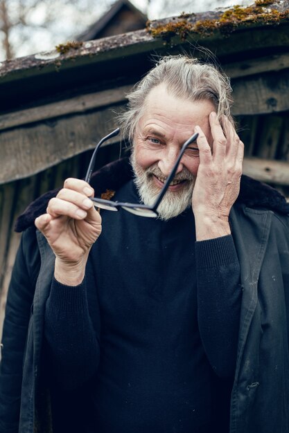 Anciano con barba se encuentra en la aldea cerca de un cobertizo de madera con un abrigo de piel de oveja y gafas