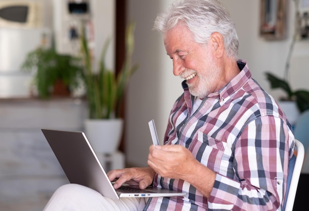 Anciano barbudo navegando por computadora portátil disfrutando de las compras en línea Alegre y sonriente hermoso anciano en casa de buen humor usando tarjeta de crédito para comprar