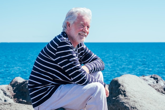 Un anciano barbudo sentado cerca del mar admirando la belleza de la naturaleza un hombre caucásico sonriente disfrutando de un día soleado y de verano en retiro o vacaciones