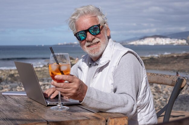 Un anciano barbudo sonriente trabaja en una computadora portátil al aire libre en la playa disfrutando de una bebida fresca