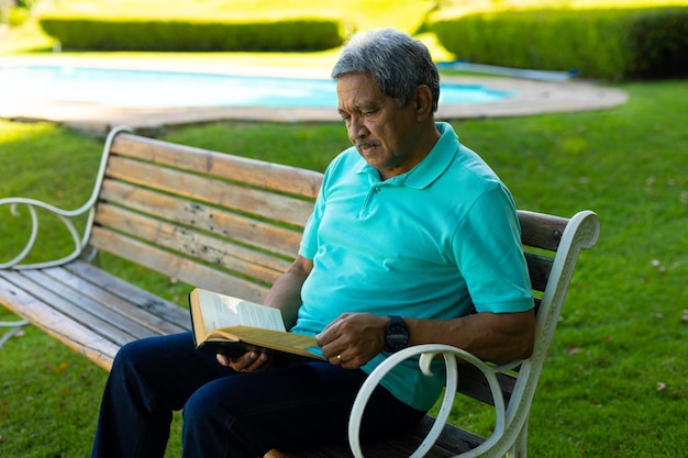 Un anciano birracial enfocado con una camiseta azul leyendo un libro mientras se sienta en un banco en el parque