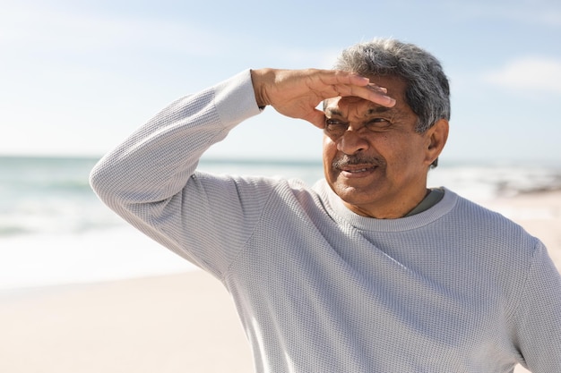 Un anciano birracial jubilado protegiéndose los ojos mientras mira a la playa durante el día soleado