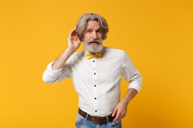 Anciano de cabello gris con bigote barbudo en camisa blanca pajarita posando aislado sobre fondo amarillo anaranjado en el estudio. Concepto de estilo de vida de las personas. Simulacros de espacio de copia. Trate de escuchar el juguete con la mano cerca de la oreja.