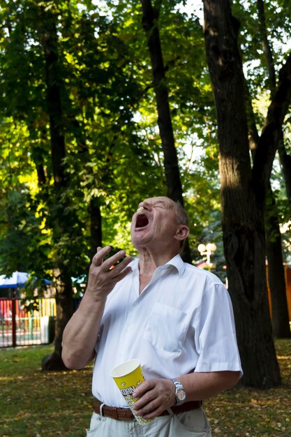 Un anciano camina solo en el parque en el verano El jubilado arroja alegremente palomitas de maíz de un vaso y las atrapa con la boca abierta