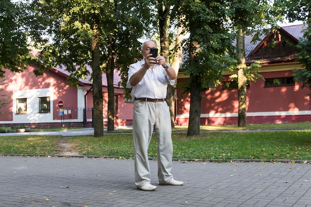 Un anciano camina solo en el parque en el verano Un moderno hombre de negocios jubilado con una camisa blanca y pantalones toma fotos con una cámara en un teléfono móvil