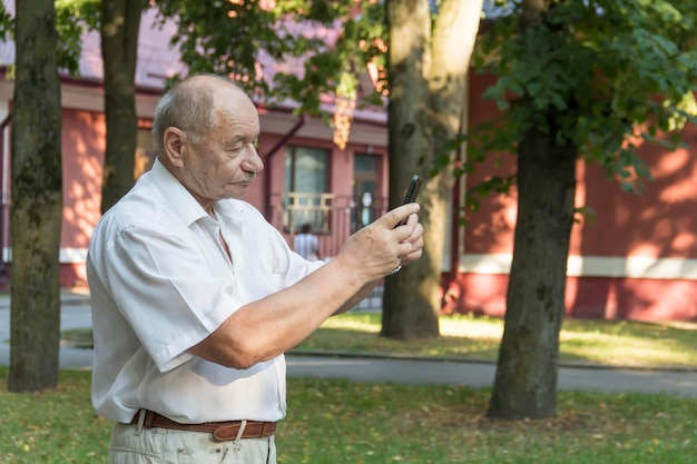 Un anciano camina solo en el parque en el verano Un moderno hombre de negocios jubilado con una camisa blanca y pantalones toma fotos con una cámara en un teléfono móvil