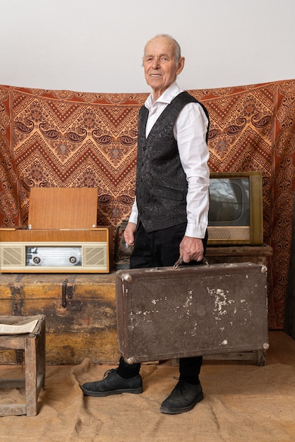 Anciano con camisa blanca sostiene el viejo equipaje clásico de maletero de viaje, se encuentra entre la sala vintage con televisión y radio antiguas