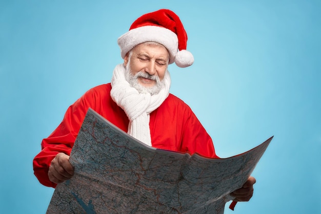 Anciano canoso con gorro rojo de Papá Noel, chaqueta y bufanda de punto mirando un gran mapa de papel y buscando direcciones para entregar los regalos de los niños. Santa Claus planeando su viaje navideño.