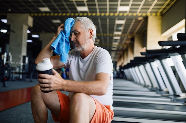 Anciano cansado con toalla bebe agua en el gimnasio