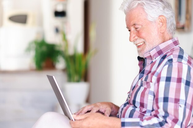 Anciano en casa navegando por una computadora portátil disfrutando de la tecnología y las redes sociales Sonriendo barbudo hermoso hombre mayor en el interior estando de buen humor navegando por la red