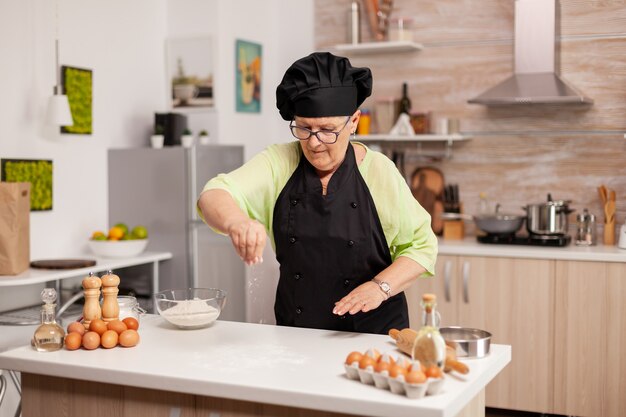 Anciano chef con uniforme rociar harina en la cocina de casa vistiendo delantal y bonette