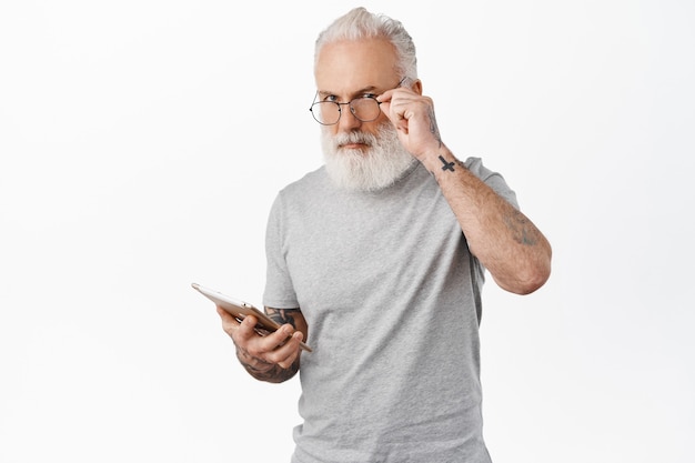 Anciano elegante con gafas con tatuajes, sosteniendo tableta digital, compras en línea con dispositivo, de pie en camiseta gris contra la pared blanca