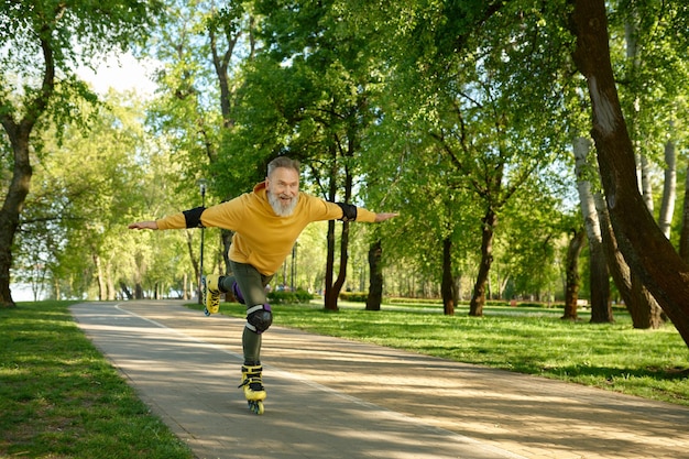 Un anciano emocional muy contento haciendo trucos mientras monta patines en un parque urbano. Varón maduro de pelo gris levantando una pierna moviéndose rápido y sintiendo satisfacción. Estilo de vida activo en la jubilación
