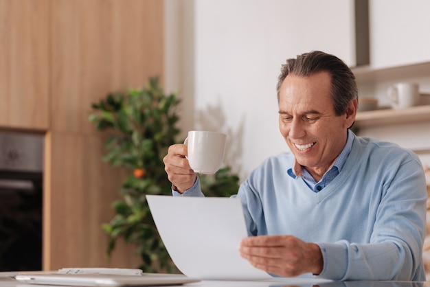 Anciano encantador positivo sentado en la cocina mientras disfruta de una taza de café y trabaja con documentos