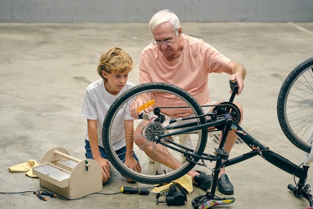 Anciano enfocado con nieto reparando rueda de bicicleta mientras está sentado en el piso de concreto