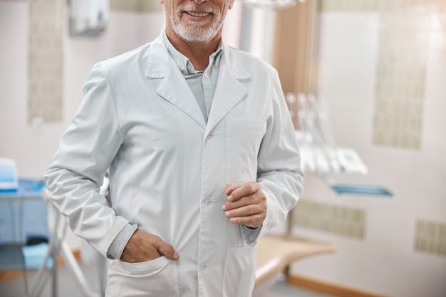 Anciano especialista dental sosteniendo su mano en el bolsillo y sonriendo en su oficina