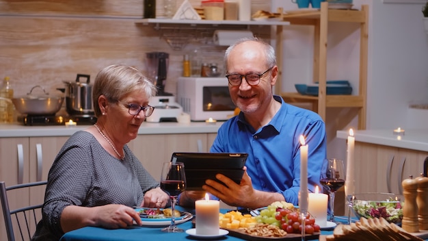 Anciano esposo jubilado senior mostrando fotos en tableta a su esposa durante la cena romántica. Pareja sentada en la mesa, navegando, hablando, usando internet, celebrando su aniversario en el comedor.